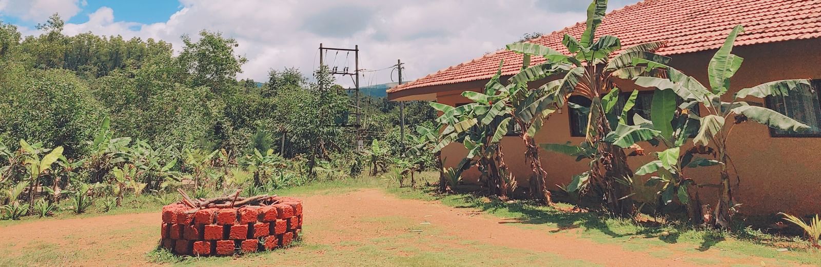 A wide outdoor view of a tiled-roof house surrounded by greenery with a circular arrangement of red-painted well in the garden at Paradise Wild Hills Resort, Kollur.