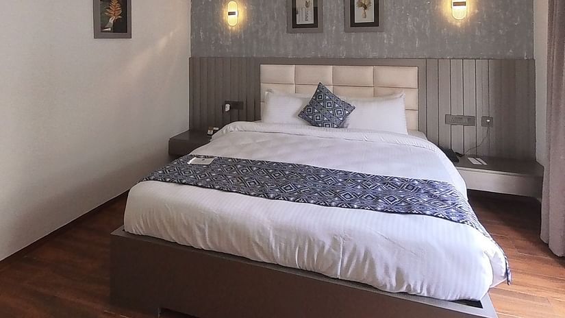 A hotel bedroom at Grand Continent, Anjuna featuring a neatly made bed, textured wall, two framed pictures, and sconce lights above the headboard. An AC unit is on the wall.