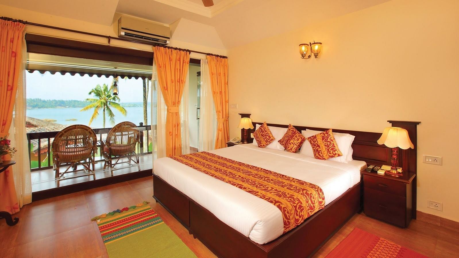 An image of Premium lake View Villa with Private Terrace featuring a double size bed, a bedside table with lamp, and a balcony attached which has two chairs placed- Fragrant Nature Kollam