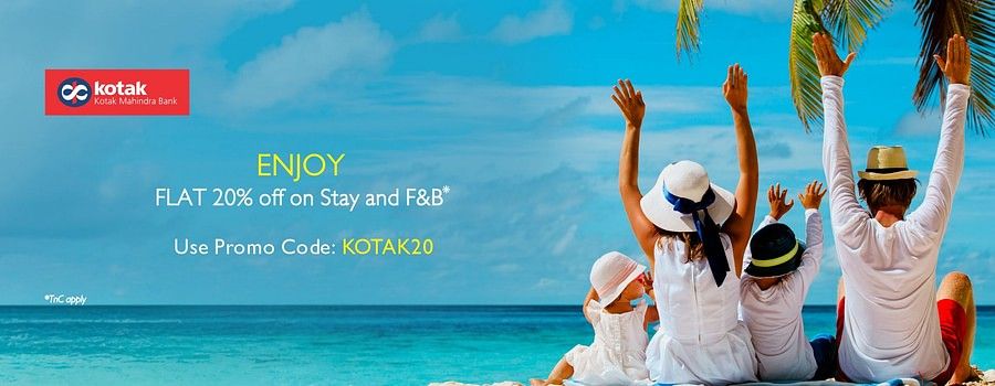 A cheerful family of four sits on a tropical beach facing the ocean, raising their hands in joy under a clear blue sky with palm trees. A promotional banner offers 20% off on stay and F&B for Kotak Mahindra Bank customers.