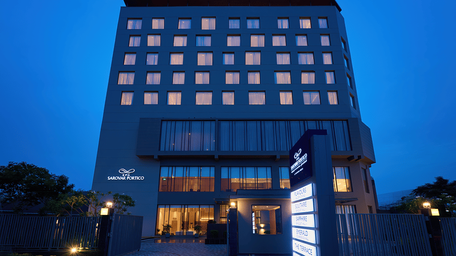 A hotel building exterior at KPG Sarovar Portico, Raj Nagar Extension, Ghaziabad featuring a multi-storey facade, illuminated entrance, driveway, and a property sign post.