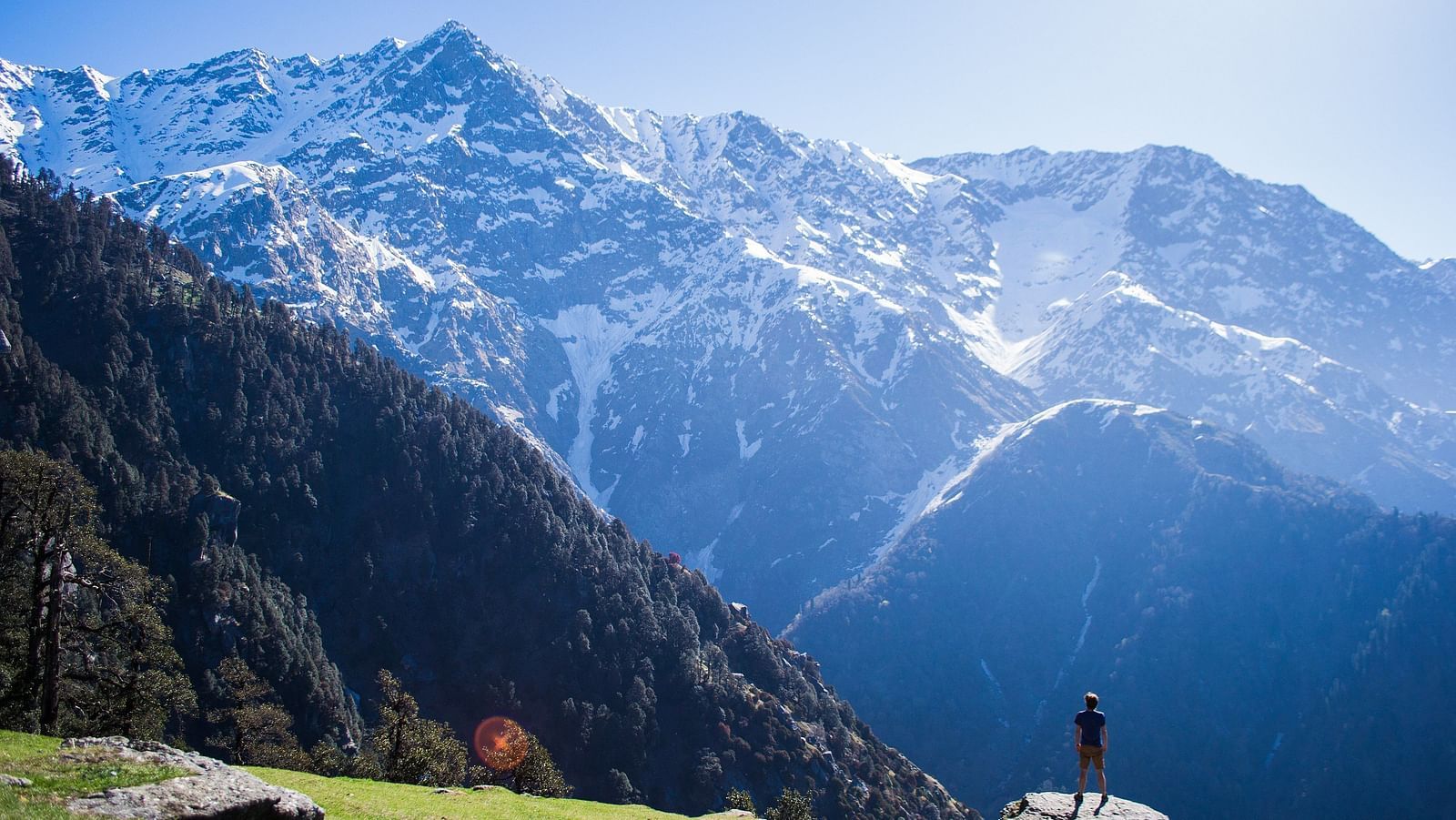 An image of snow caped mountains where a person is seen standing near a cliff enjoying the view