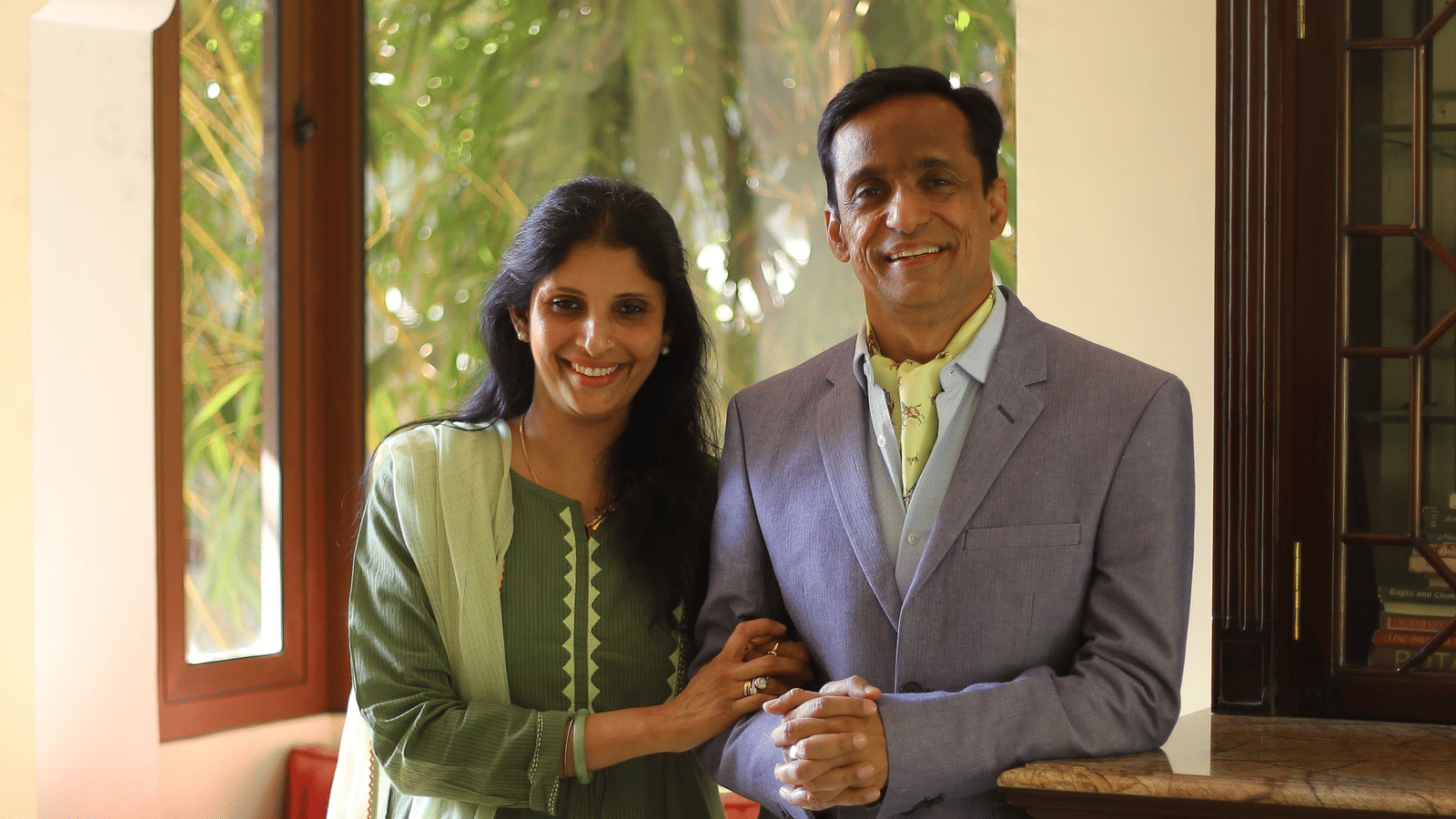 Mr. Anirudh and his wife Mandvi standing together indoors, smiling at the camera, with greenery visible through the window and warm seating and wooden furnishings in the background.