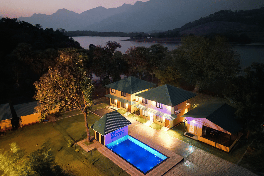 High-angle night view of the resort showing the glowing blue swimming pool, lit pathways, and the dark silhouette of the lake and mountains - Ibex Resorts, Malampuzha (Kava Eco Camp and Caravan Park)
