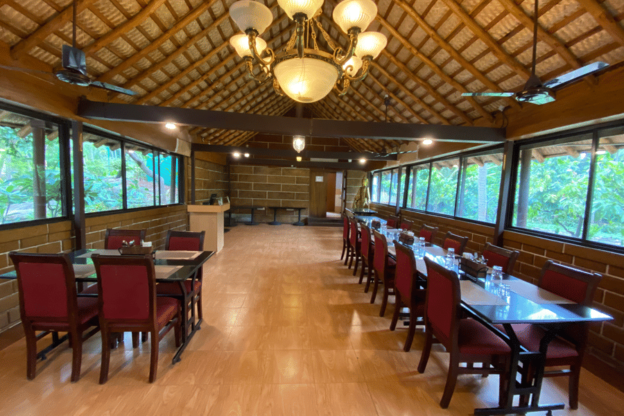 Formal dining setup at Ibex River Resort, Pollachi showcasing wooden tables and chairs, elegant chandeliers, and large windows providing natural light and scenic forested views for guests.