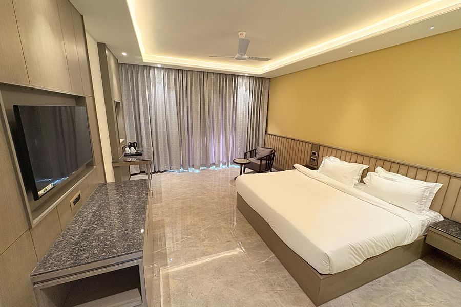 An image of a bedroom featuring double size bed, a chair with a small centre table, a flat TV screen, and a tea and coffee maker - Pride Hotels Group