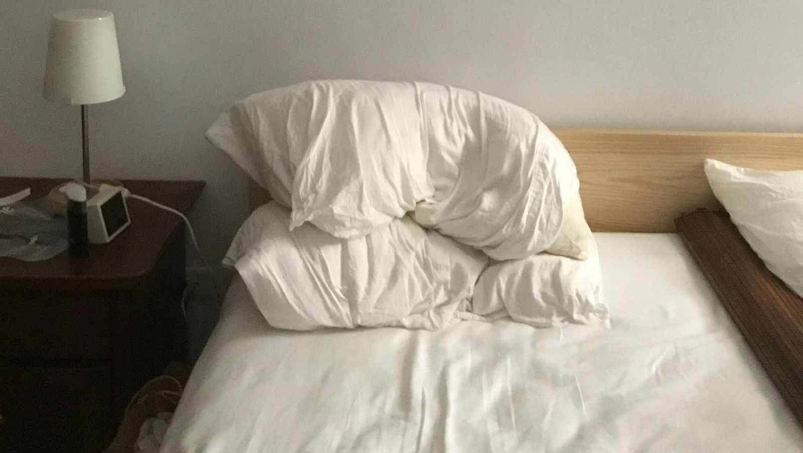Minimalist bedroom with a double bed made up with white linens and a small bedside lamp against a plain wall.
