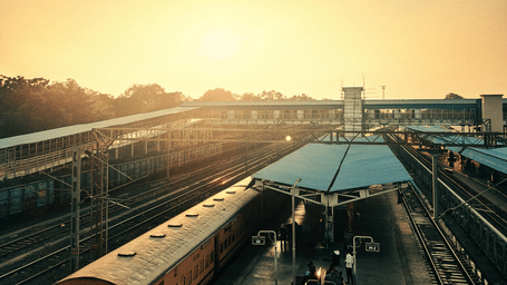 A high-angle view of a bustling Indian railway station platform with a passenger train arriving, showing overhead walkways and tracks under a bright morning sky.