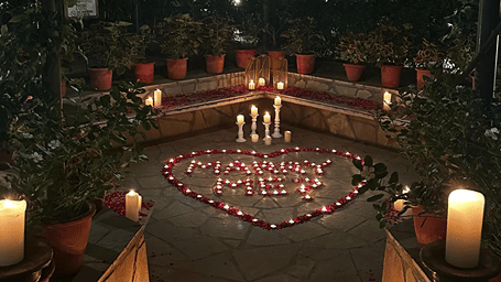 An outdoor garden area decorated with candles and flower petals forming a heart-shaped arrangement around a message, surrounded by potted plants and lanterns.