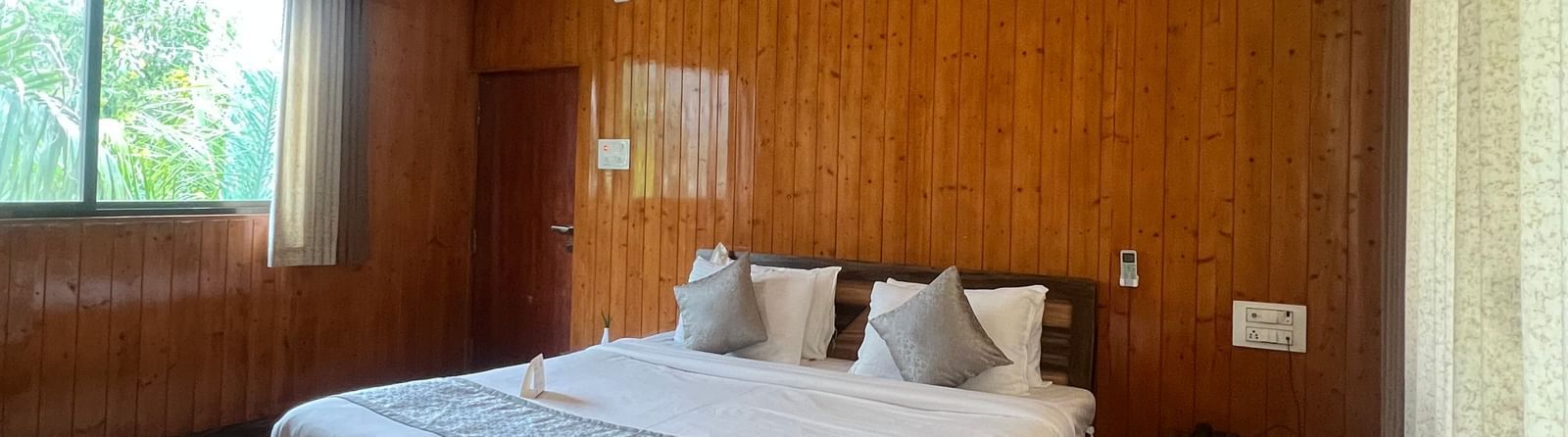 Wide view of the Daksh Elite Cottage, highlighting its wooden interiors, a window with curtains opened, and its centrally placed double bed, at Daksh The Valley Resort, Saputara.