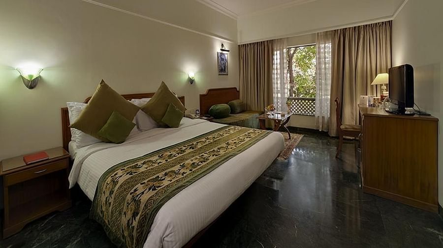 A comfortable hotel room at Renest Shraddha Inn features a bed with decorative pillows and a patterned runner, a seating area, a television on a stand, and large windows with curtains - Renest Shraddha Inn, Shirdi