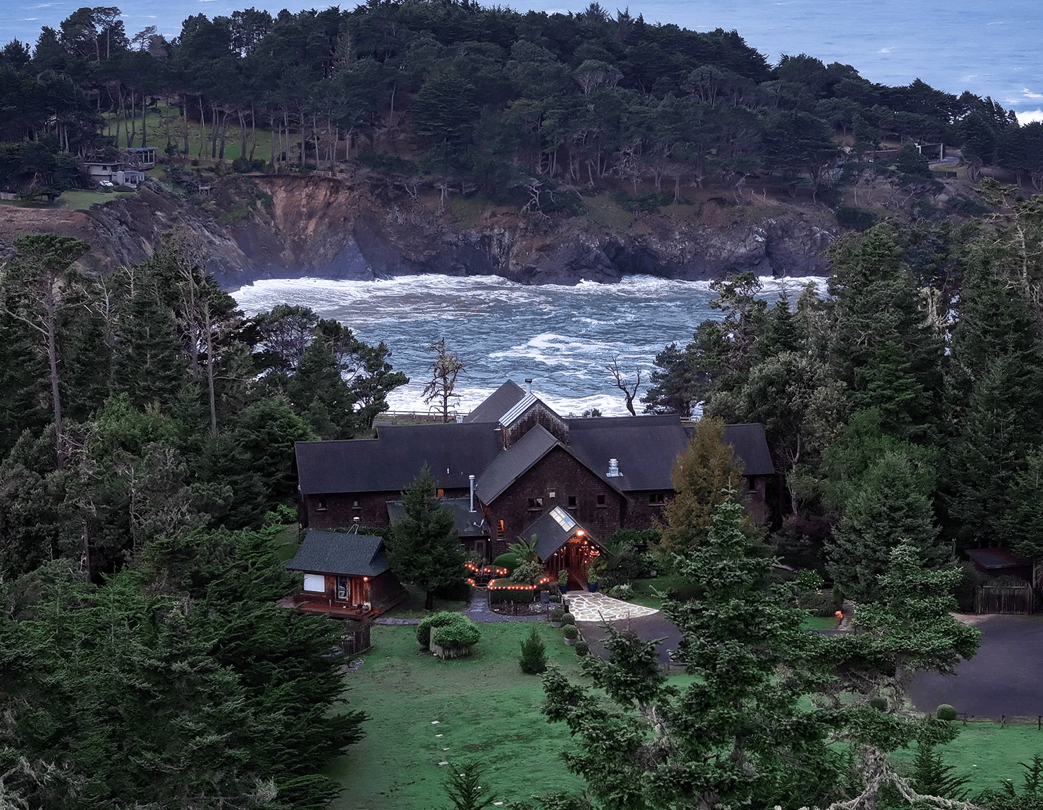 A house nestled among tall trees on a coastal cliff overlooking a turbulent ocean under an overcast sky in the background at Brewery Gulch Inn.