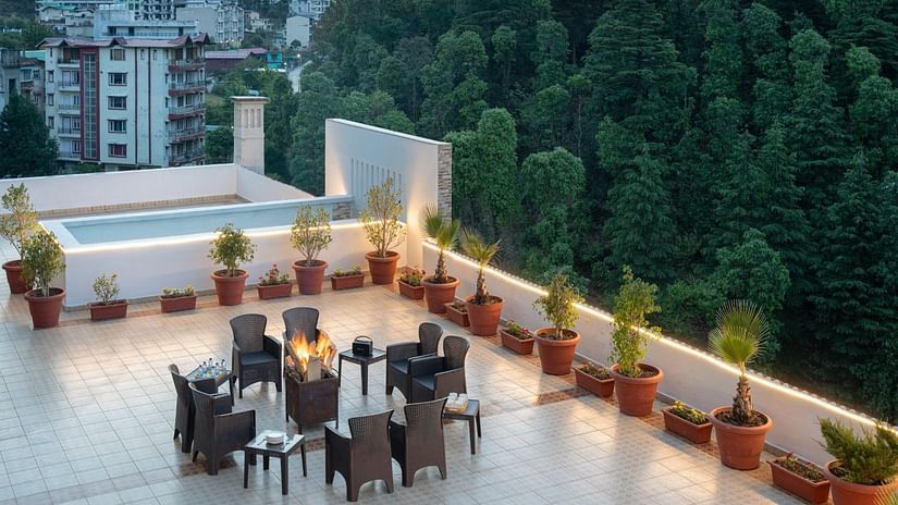 A rooftop terrace features a fire pit surrounded by chairs, potted plants, a shallow pool, and a view of buildings and lush greenery at Clarks Inn, Near Kainchi Dham.