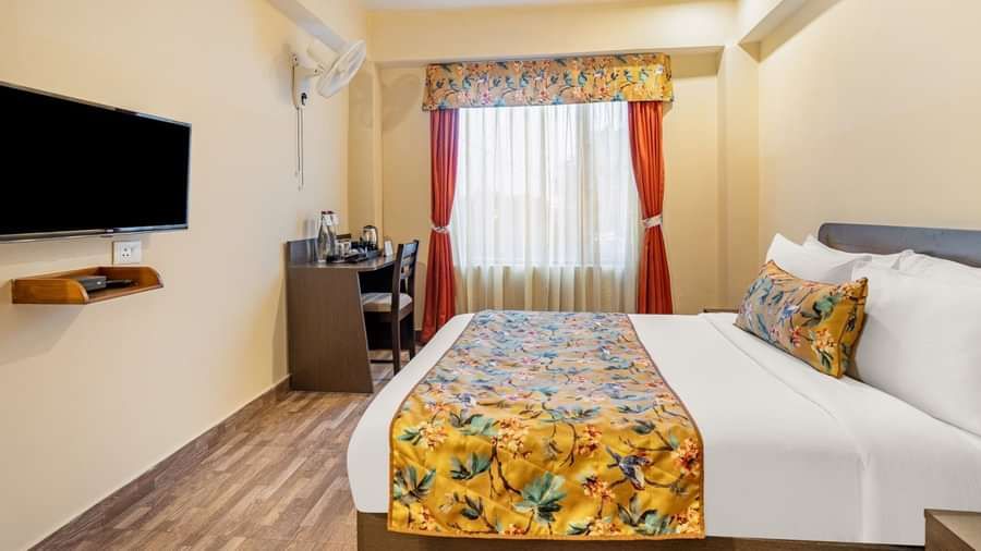 Bright hotel room with a comfortable double bed, floral bedding, flat-screen TV, work desk with water bottles and kettle, and a large window with orange curtains - Summit Namnang Courtyard & Spa, Gangtok