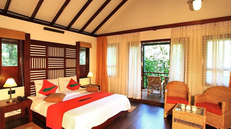A bed placed in a room with two chairs and table, and a balcony that offers forest views | Vythiri Resort, Wayanad