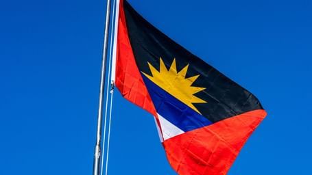 The flag of Antigua and Barbuda, featuring a black triangle with a rising yellow sun over a blue band, flanked by red triangles on either side, flying against a clear blue sky.