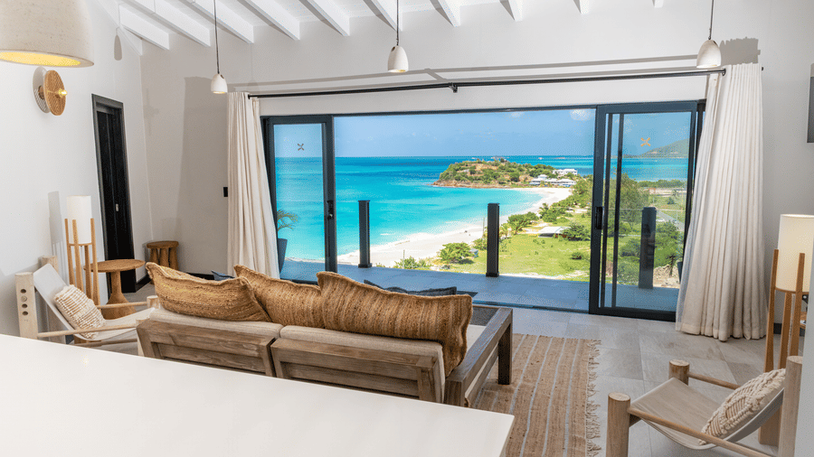 A living area with a sofa, two lounge chairs, pendant lights and ocean views through large windows at Tamarind Hills Resort and Villas, Antigua.