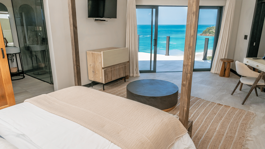 A bedroom with a king size bed, a cabinet, a table, a chair and floor-to-ceiling windows offering panoramic ocean views at Tamarind Hills Resort and Villas, Antigua.