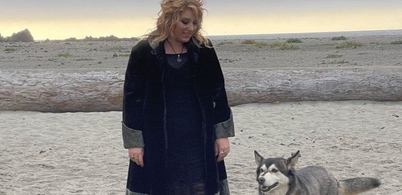 A person in a coat and hat standing on a beach with a dog.