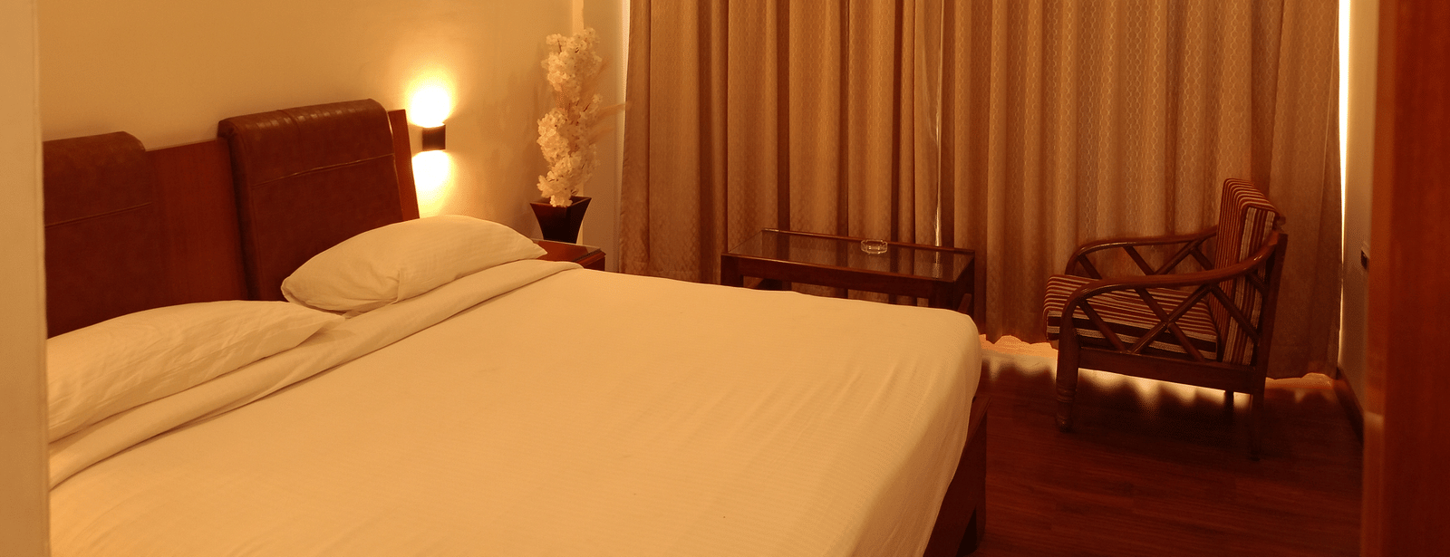 Guest room at Hotel Gold, Panipat featuring a bed, chair, side tables, and large window curtains.