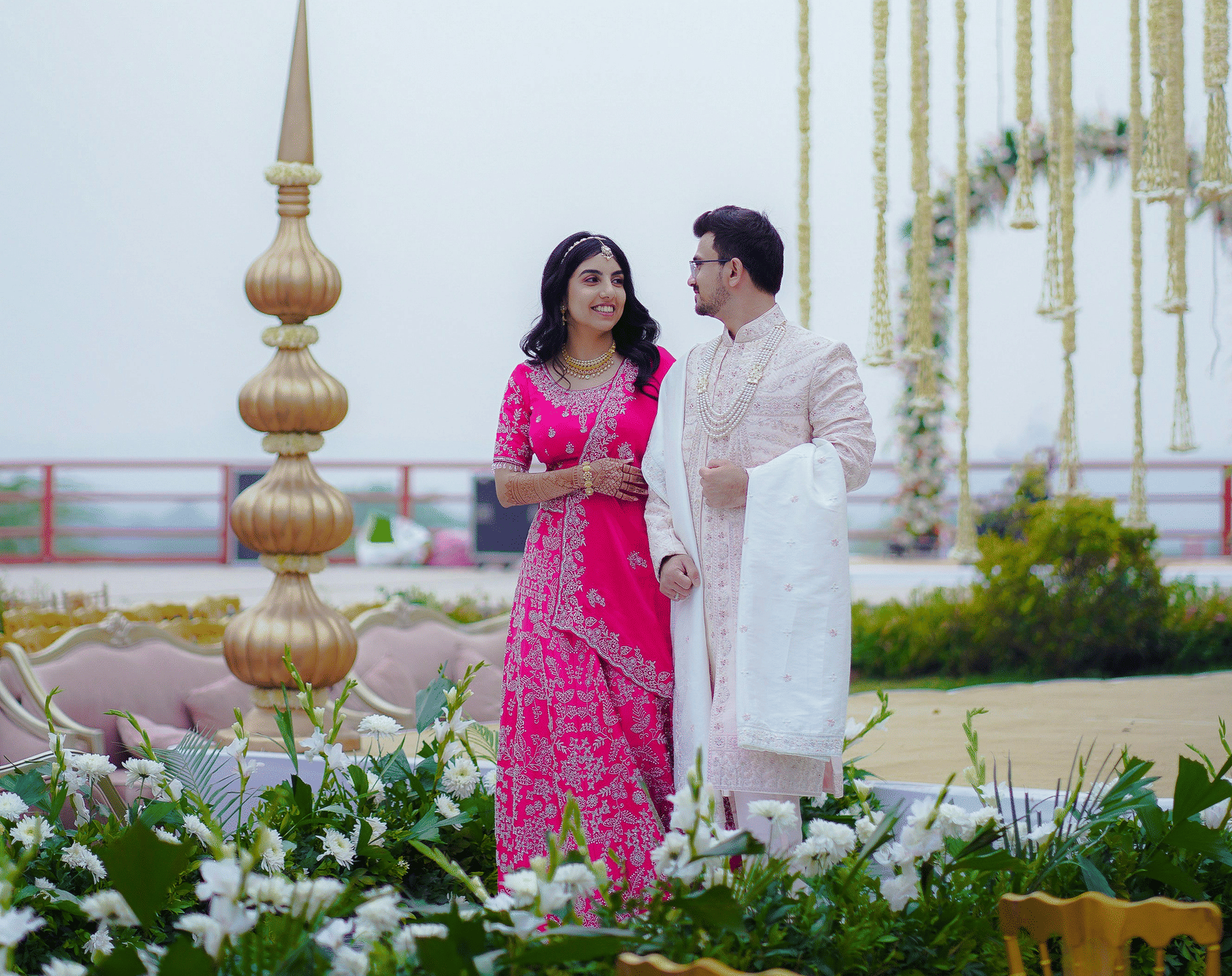 A bride and groom in pink and dark clothing stand in a light garden setting, leaning toward each other by a white floral archway.