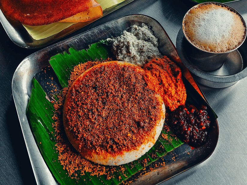 A food spread of thatte idli, filter coffee, and doas served on steel plates with banana leaves with chutney and sambar.