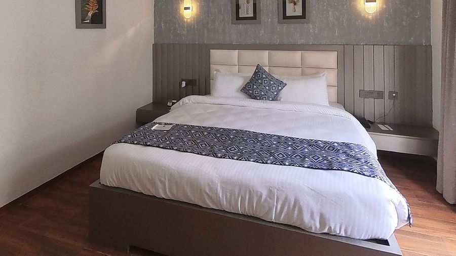 A hotel bedroom at Grand Continent, Anjuna featuring a neatly made bed, textured wall, two framed pictures, and sconce lights above the headboard. An AC unit is on the wall.