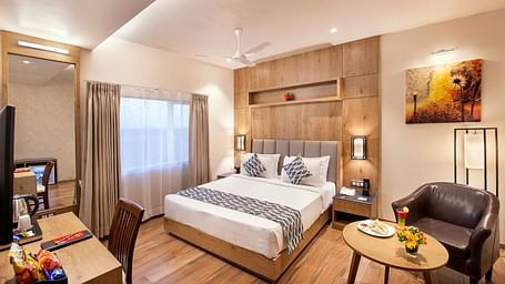 A hotel room showcasing a double bed with white and grey bedding, a wooden bedside table with a lamp, a work desk area with a chair and television, and a small round table with a brown armchair, complemented by wooden flooring at Lords Inn Motikhavdi