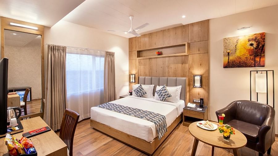 A hotel room showcasing a double bed with white and grey bedding, a wooden bedside table with a lamp, a work desk area with a chair and television, and a small round table with a brown armchair, complemented by wooden flooring at Lords Inn Motikhavdi
