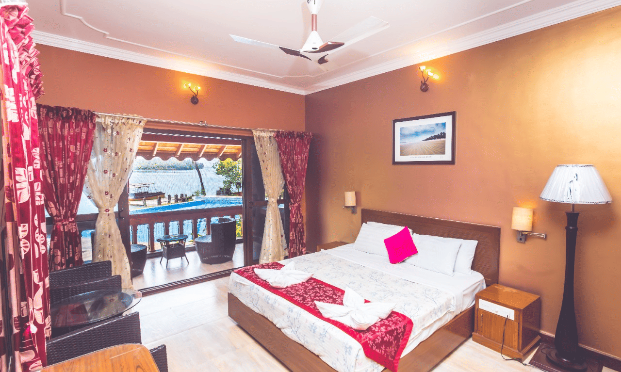 A brightly lit room with a queen bed and pink floral accents, wall art and a window overlooking the water at Paradise Lagoon Resort, Udupi.