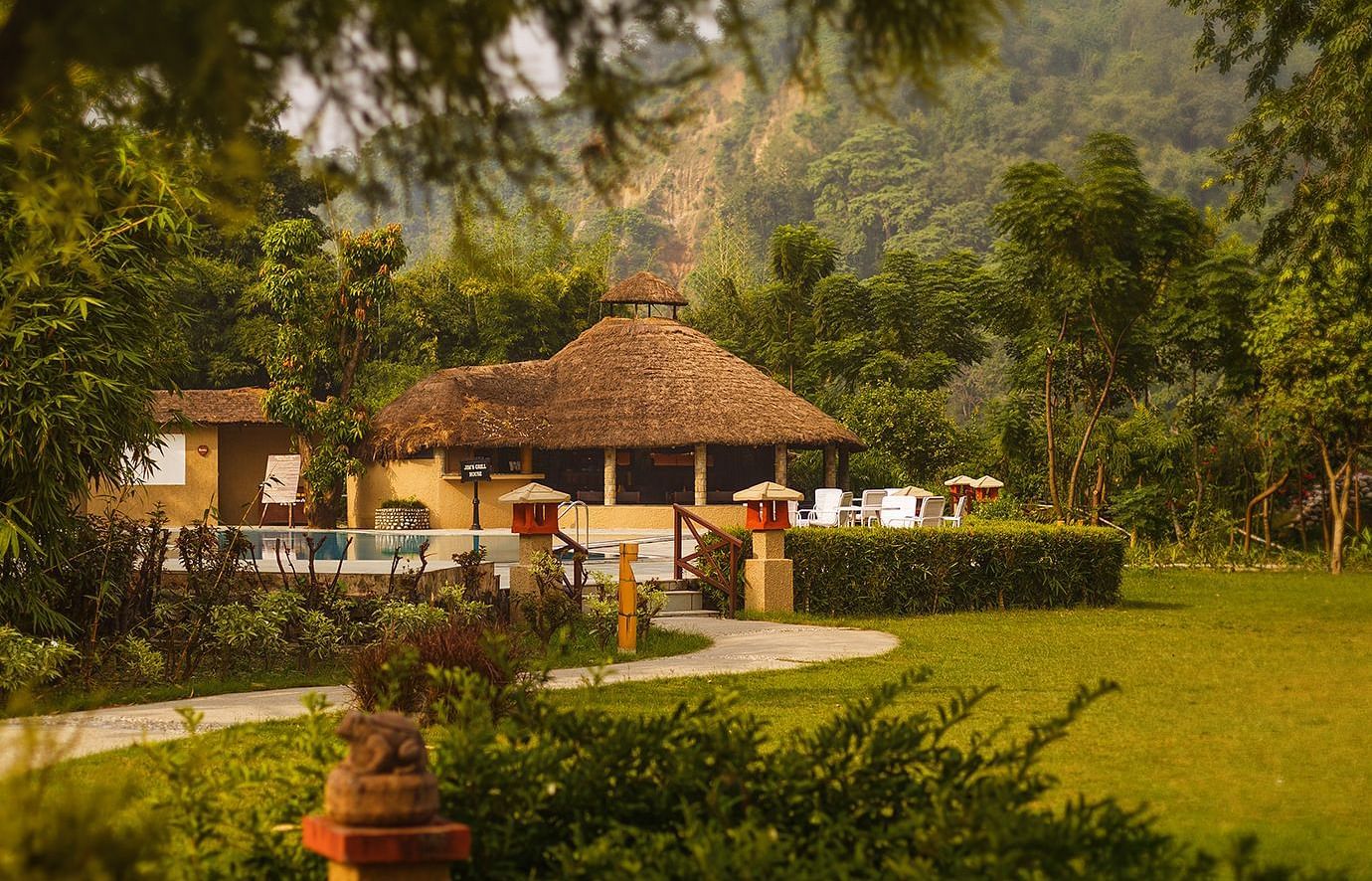 An image of a hut amidst the garden, behind it there is a outdoor seating area with chairs and tea table- The Riverview Retreat, Corbett