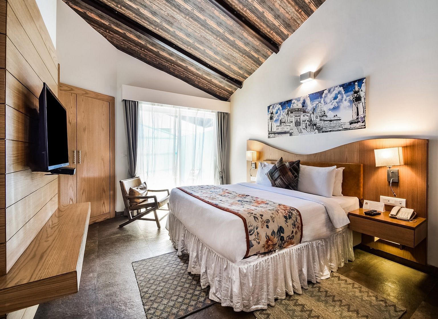 A cosy hotel room with wooden walls, a Tv mounted on the wall, a bed with pillows on it and a balcony.- Bamboo Saa Resort & Spa, Udaipur