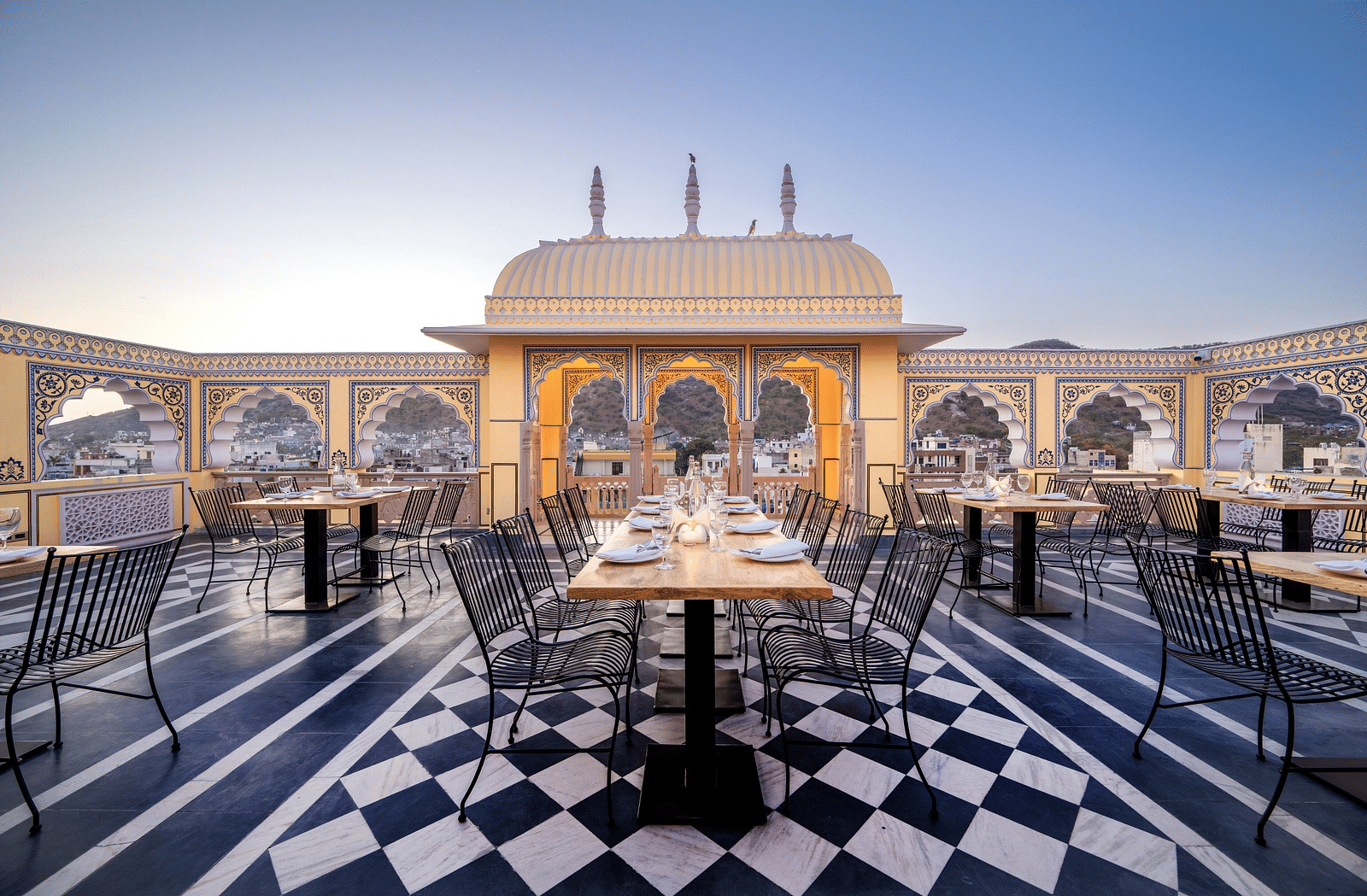 a close up image of a long dining table at our terrace lounge with a balcony adorned with rajasthani artistry behind it - Brij Castle, Jaipur