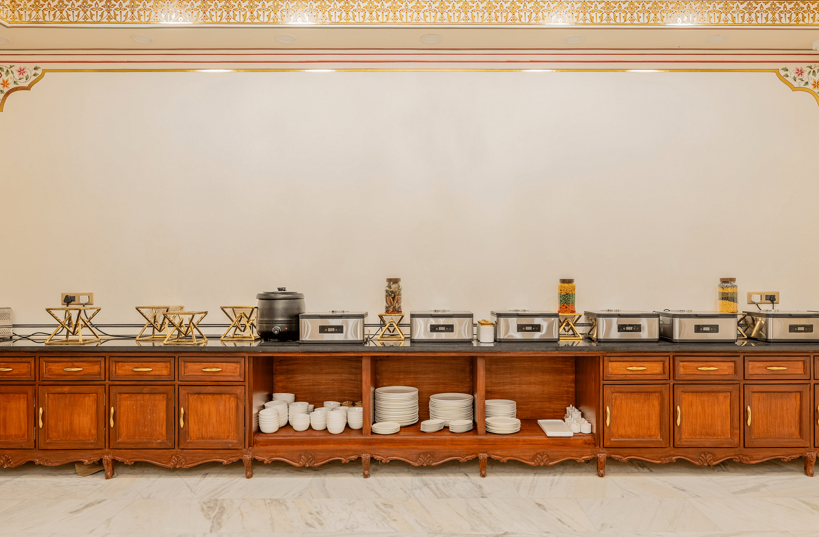 a buffet counter with well stacked plates and bowls and other cutlery in the self below - Brij Castle, Jaipur