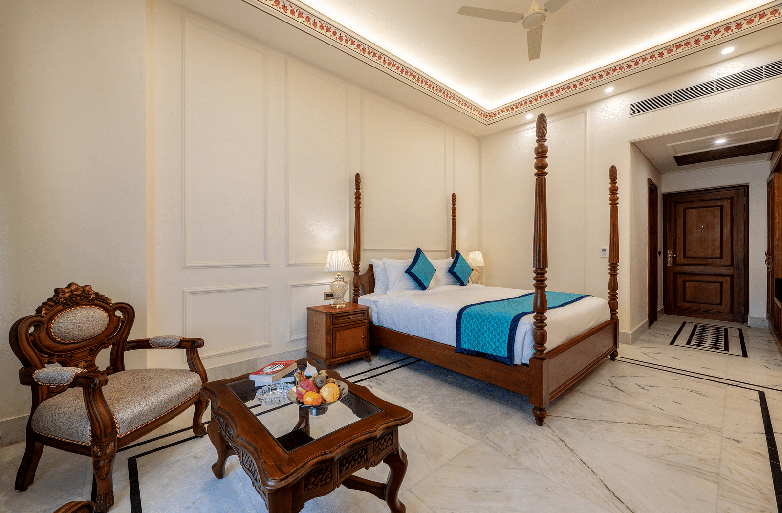 an arm chair with a coffee table in front of it featuring a bowl of fruits along with a plush king size bed behind it - Brij Castle, Jaipur