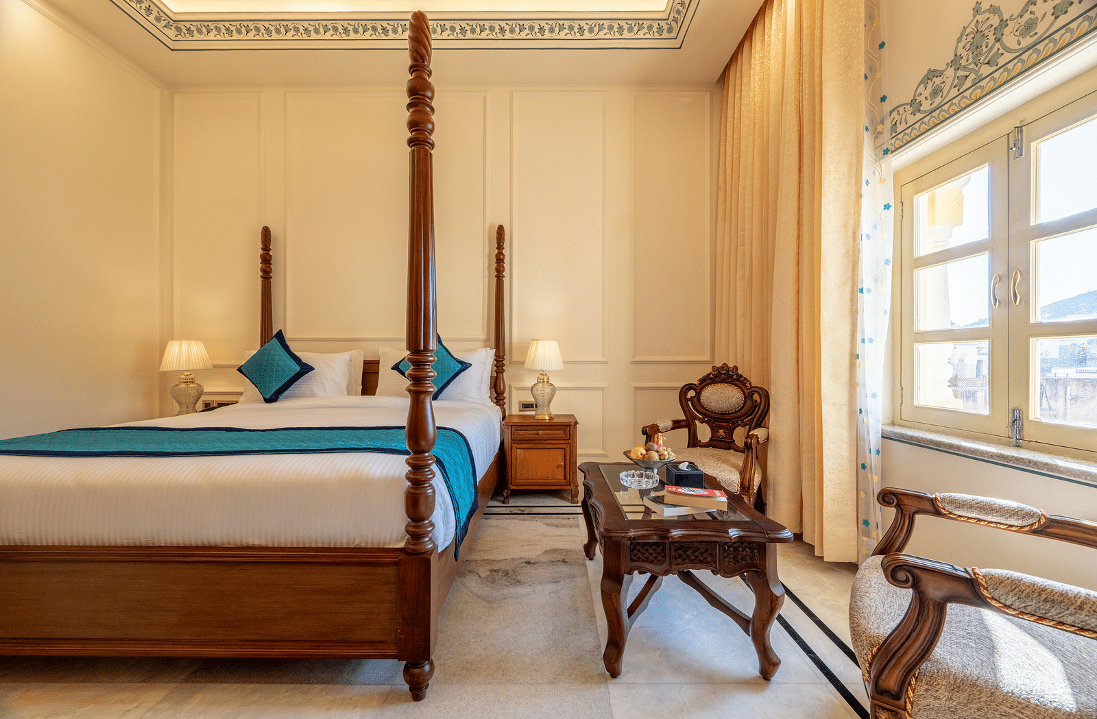 a bedroom featuring a king size bed along with two arm chairs placed in front of a coffee table with a decorative wooden frame behind it - Brij Castle, Jaipur