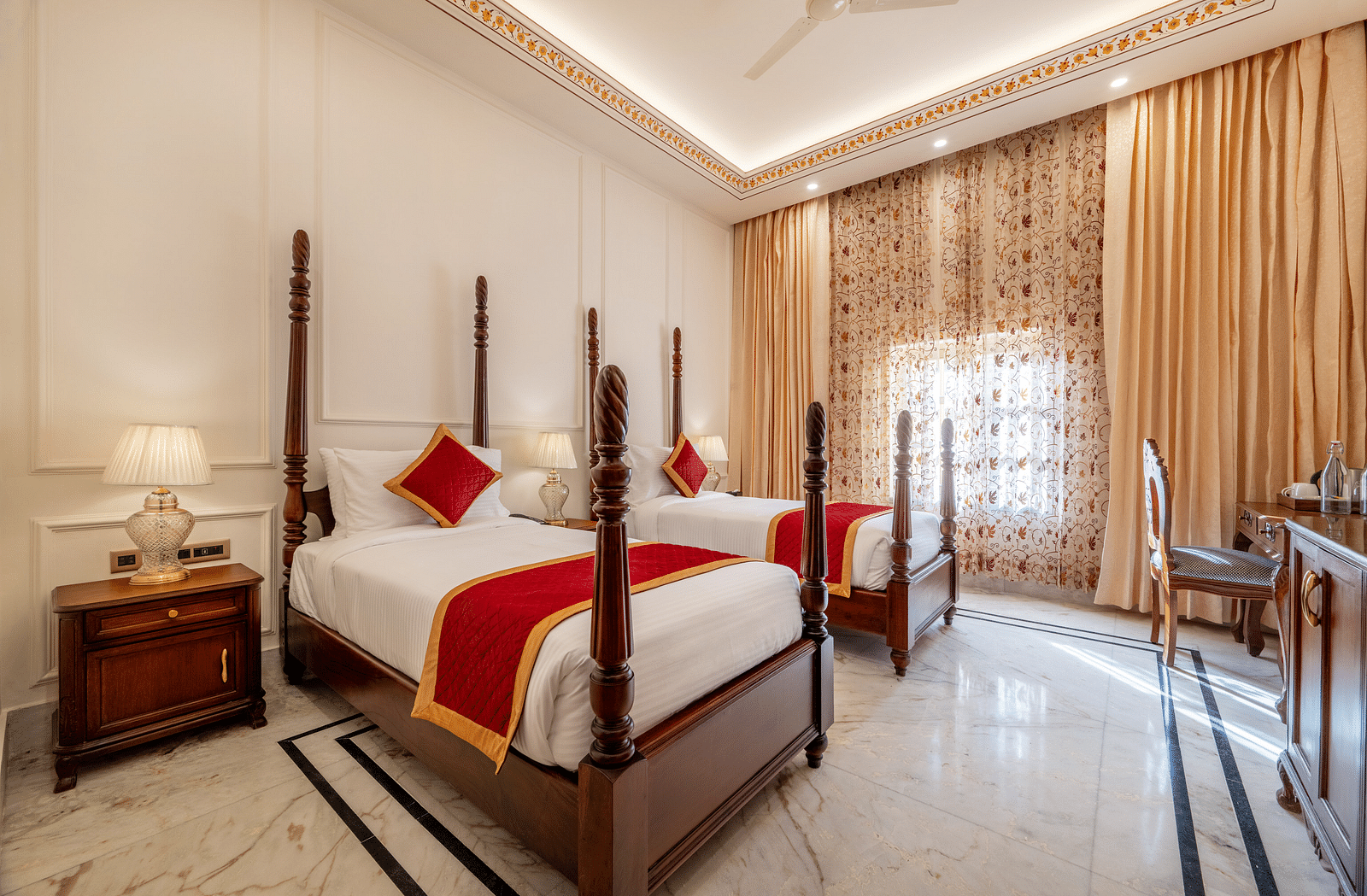 corner view image of a twin bed room along with a work desk and chair with tall golden curtains covering the window - Brij Castle, Jaipur
