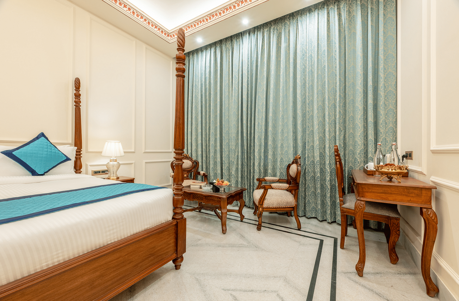 a brightly lit room with teal coloured curtains covering the windows with a double bed and a work desk - Brij Castle, Jaipur