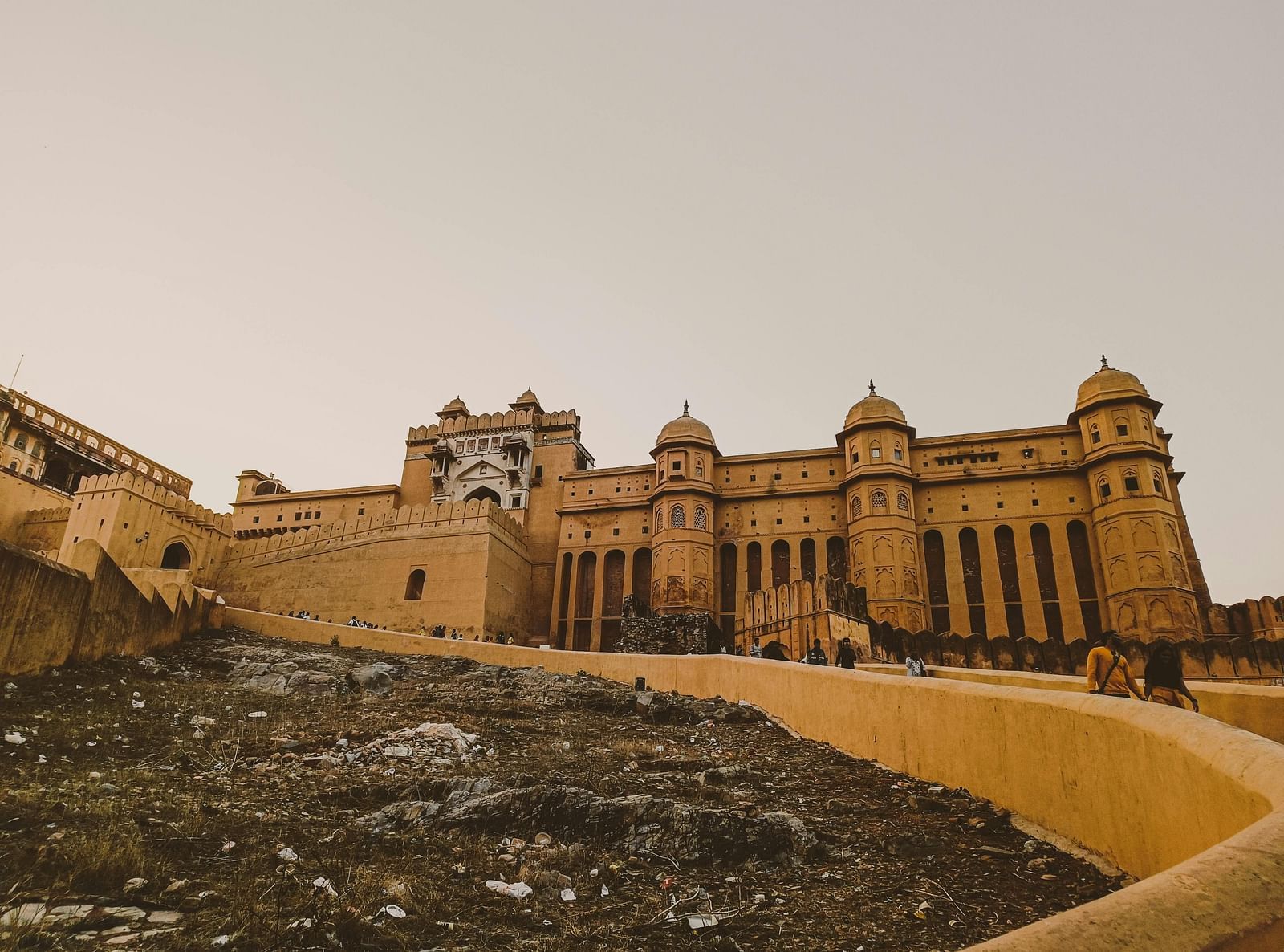 A wide shot of Amber Fort with yellow sandstone walls and ramparts against a pale sky, with a rough foreground