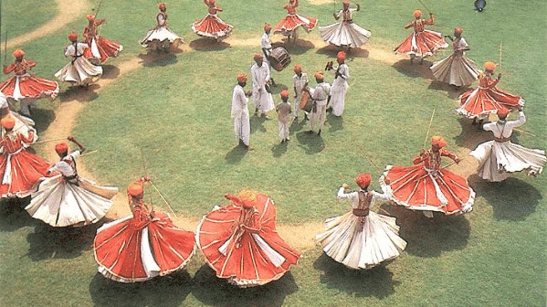 Dancers in traditional red and white attire whirl in a large circle on a green field, performing a vibrant folk dance - Umaid Palace