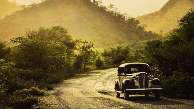 Vintage car winding through a misty forested road with sunlit hills in the background.