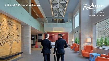 Creative corporate photography showing two businessmen in suits walking through the modern, spacious lobby of Radisson Bengaluru City Center Ulsoor Lake, featuring contemporary design elements and orange accent furniture, promoting the Business Class Room 