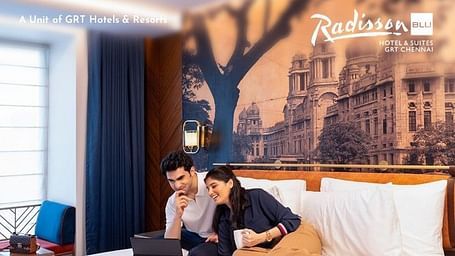 Creative hotel photography featuring a romantic couple relaxing together on a plush hotel bed while using a laptop, with Chennai's heritage architecture visible through the window, highlighting the cozy Independence Day weekend staycation experience.