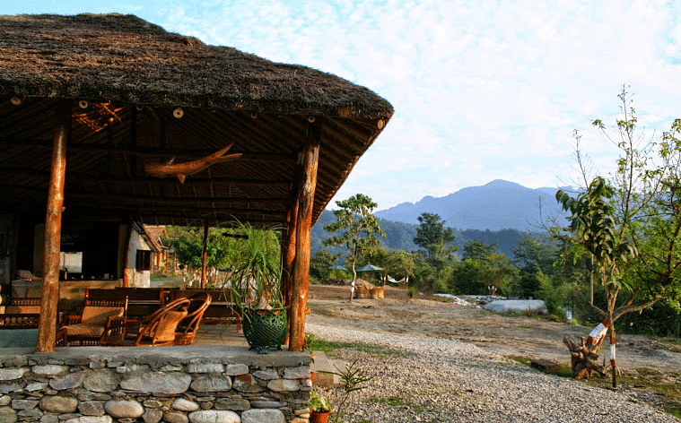 image shows a thatched-roof tent stay at the jamoon corbett, with a view of the mountains in the background.