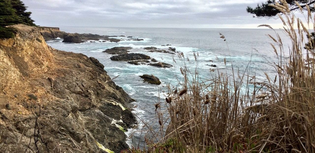A rugged coastline with waves crashing against the rocky cliffs under a cloudy sky.