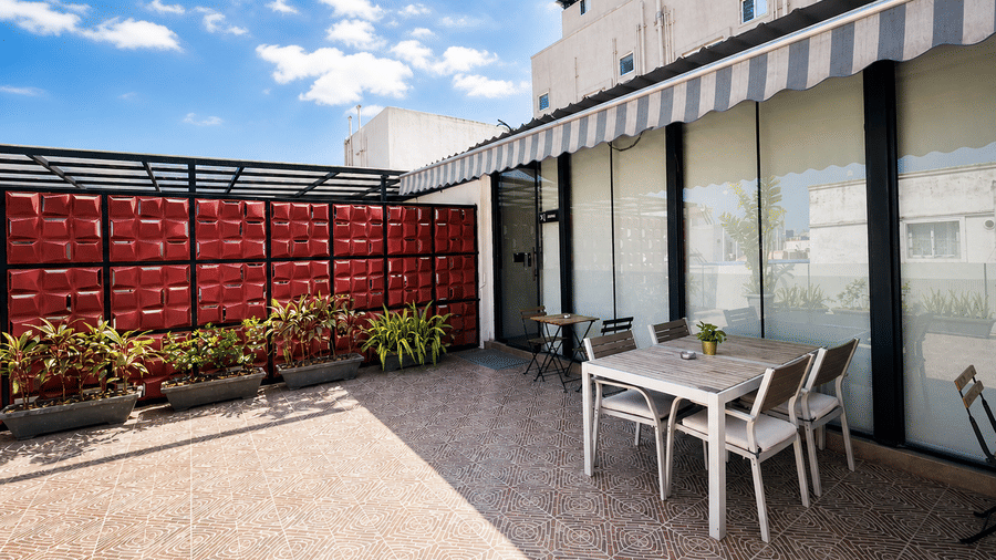 Open rooftop terrace at Hotel Kyzen Smart Stays Madhapur with white chairs and tables, red boundary walls, potted plants, and a striped canopy overhead.