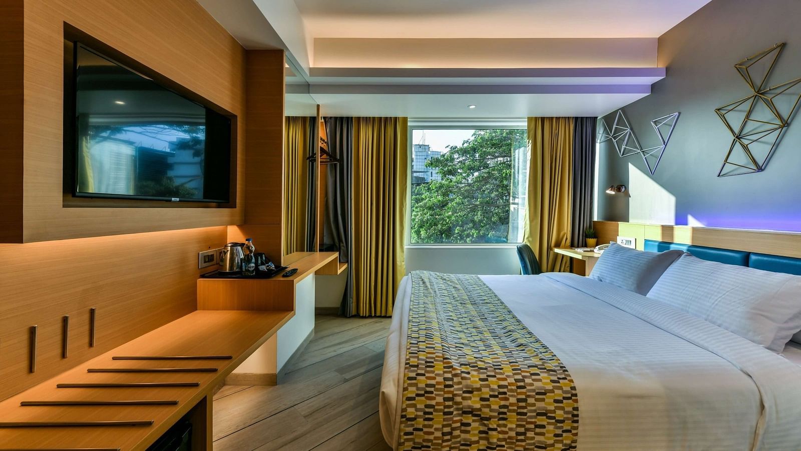 A spacious room featuring a large bed and a window with curtains pulled to the side, offering a view of trees, at Indie Stays, BKC Kalina.