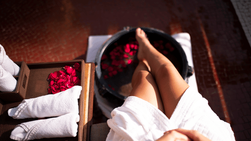 An image of a woman's leg resting in a round tub filled with water and rose petals, with towels neatly arranged on a tray beside it.