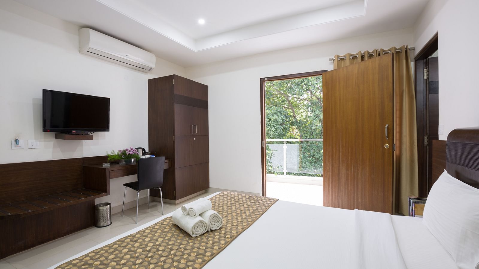 A room with a double bed, a work desk and a chair, wardrobe, and an open door to the balcony - Sanctum Suites BEL Road, Bangalore