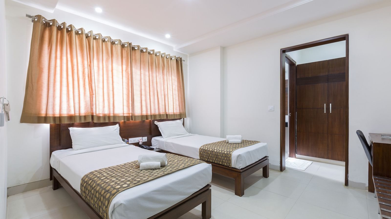 A room with twin beds featuring patterned bed runners and curtains, with an open doorway - Sanctum Suites BEL Road, Bangalore